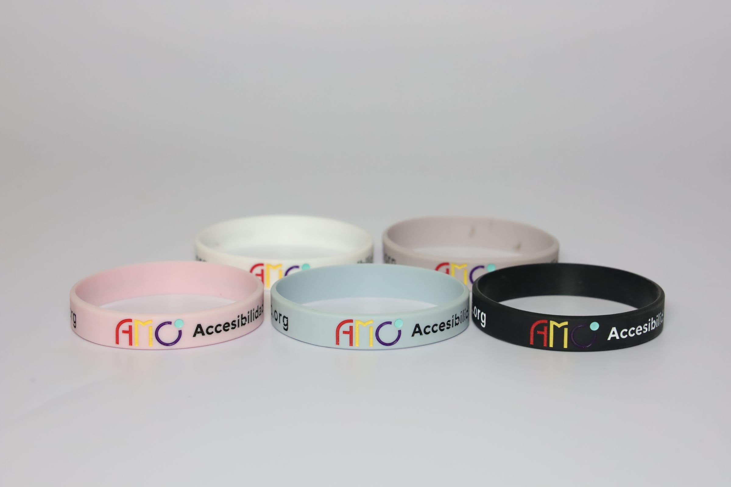 Las cinco pulseras de AMO sobre fondo gris claro: rosa, lila, negra, blanca y gris, con el logotipo y el texto Accesibilidad en Movimiento